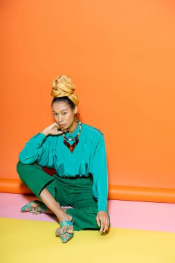 Parlak kıyafetli modacı Afro-Amerikan modeli renkli arka planda otururken kameraya bakıyor. 