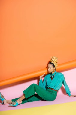 Parlak bluz ve başörtüsü içinde renkli arka planda oturan modaya uygun Afro-Amerikan modeli. 
