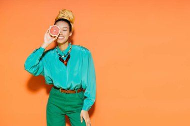 Turuncu arka planda greyfurt tutan, yaz kıyafeti içinde gülümseyen Afro-Amerikan modeli. 