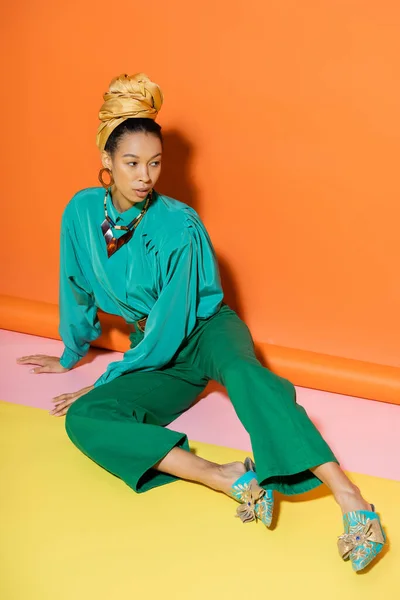 Trendy african american model in blouse and pants sitting on colorful background