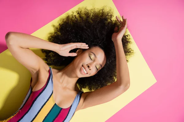 黄色とピンクの背景に横たわるカラフルな縞模様の水着でアフリカ系アメリカ人モデルの喜びのトップビュー — ストック写真