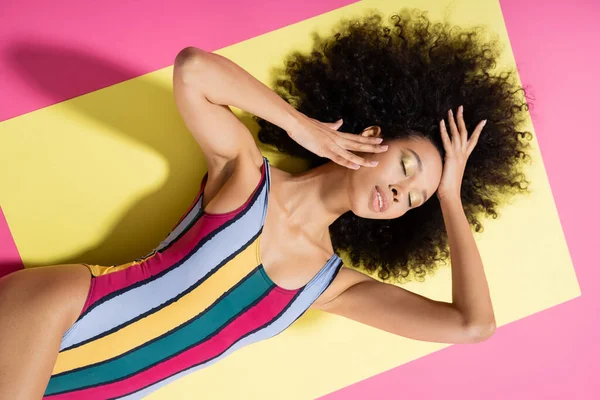 黄色とピンクの背景に横たわるカラフルな縞模様の水着で屈託のないアフリカ系アメリカ人モデルのトップビュー — ストック写真