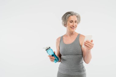 happy senior woman in sportswear holding sports bottle with water and using smartphone isolated on grey  clipart