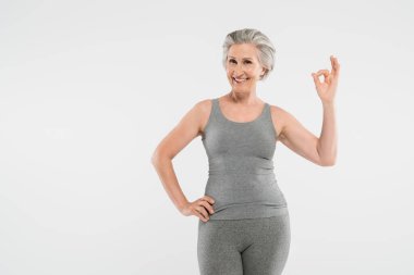 Spor kıyafetleri içinde mutlu ve emekli bir kadın. Elleri kalçalarında poz veriyor. Gri renkte izole edilmiş bir şekilde iyi görünüyor.