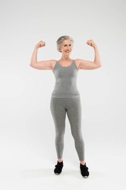 full length of joyful and retired woman in sportswear showing muscles on grey  clipart