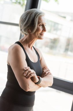 Spor salonunda kolları çapraz dururken kolları bağlı gülümseyen atletli neşeli yaşlı bayan. 