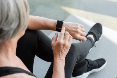 top view of senior woman with grey hair sitting and using fitness tracker after working out in gym  clipart