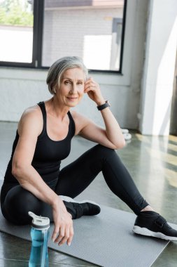 Spor giyim bölümünde son sınıf öğrencisi müzik dinliyor ve spor salonundaki spor minderinde oturuyor. 