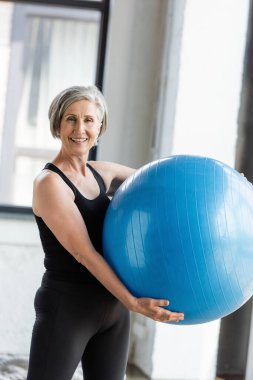 Spor kıyafetleri içinde spor salonunda spor yaparken elinde mavi fitness topu tutan mutlu son sınıf kadını. 
