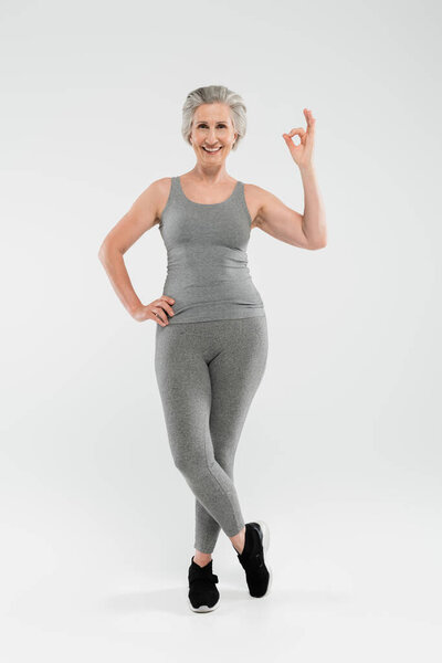 full length of cheerful and retired woman in sportswear posing with hand on hip and showing okay on grey 