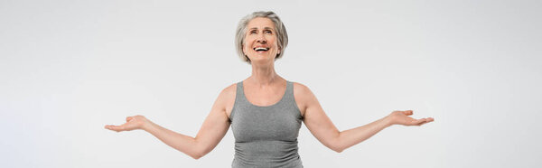 satisfied senior woman with grey hair looking up and gesturing isolated on grey, banner 