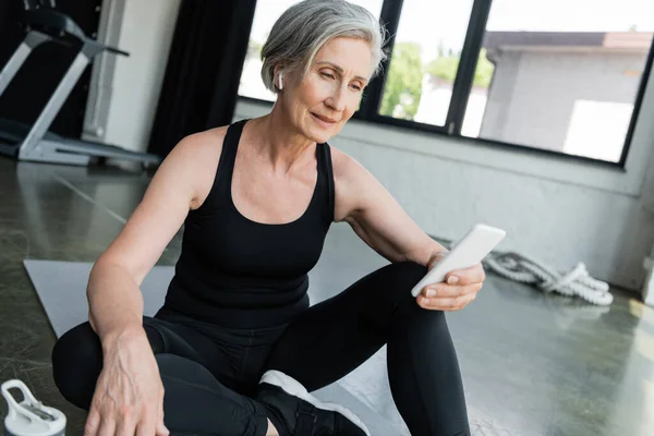 stock image senior sportswoman using mobile phone and sitting on fitness mat in gym 