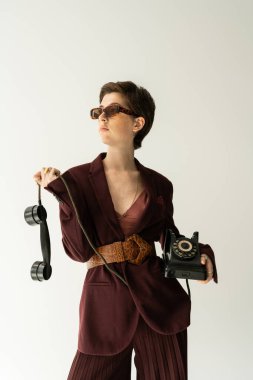 Esmer, güneş gözlüklü, bordo kahverengi ceketli, deri kemerli, gri renkli telefonlu bir kadın.