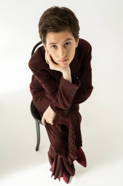 Gri arka planda oturan düşünceli, takım elbiseli bir kadının kameraya baktığı yüksek açılı manzara.