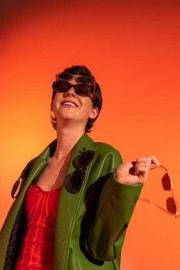excited and fashionable woman in green leather jacket posing with different sunglasses on orange background clipart