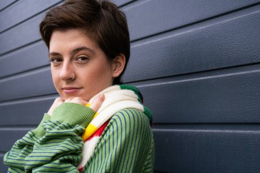 portrait of smiling freckled woman in green jumper and striped scarf standing near grey wall on street clipart
