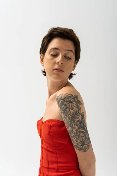 young and charming woman with tattoo posing in red corset dress isolated on grey