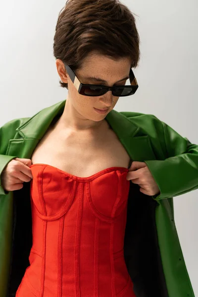 サングラスと緑の革のジャケットのスタイリッシュな女性はグレーに隔離された赤いストラップレスのドレスを調整 — ストック写真