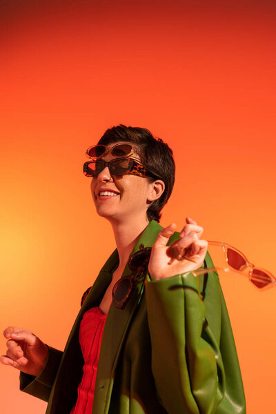 молодая и беззаботная женщина позирует в зеленой стильной куртке и различных модных солнцезащитных очках на оранжевом фоне