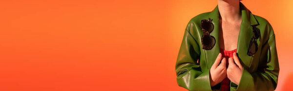 обрезанный вид женщины, позирующей в модных солнцезащитных очках на зеленой кожаной куртке на оранжевом фоне, баннер