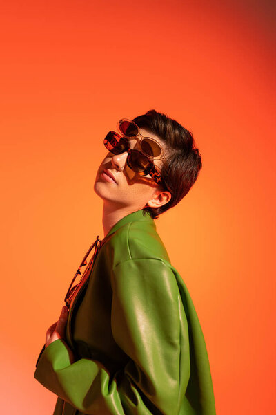 стильная брюнетка позирует в нескольких солнцезащитных очках и зеленой кожаной куртке на оранжевом фоне