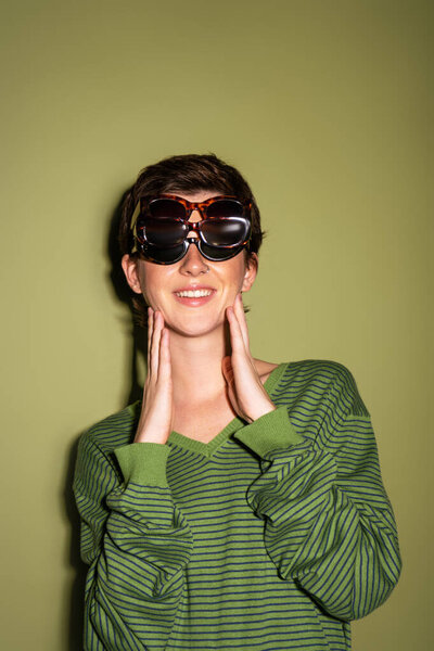 молодая и веселая женщина позирует в полосатом свитере и нескольких солнцезащитных очках на зеленом фоне