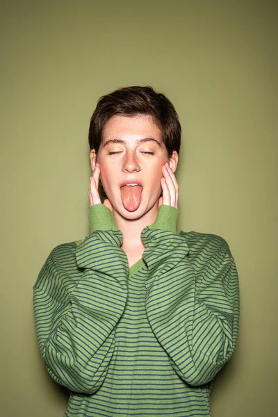 緑の背景に目を閉じて立っている間に舌を突き出す流行のジャンパーの若いモデル — ストック写真