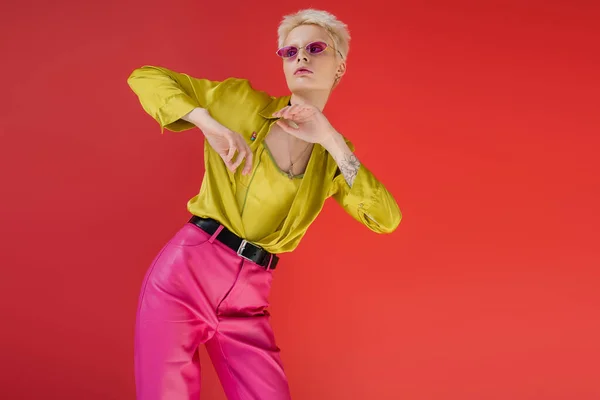 albino model with tattoo posing in stylish blouse and trendy sunglasses on carmine pink background