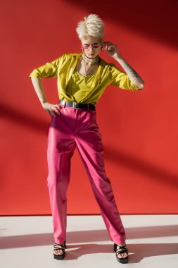 Carmine pembe arka planında eli kalçasında duran, dövmeli albino kadın. 