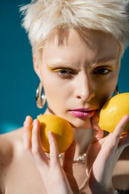 portrait of albino woman with trendy makeup and blonde hair posing with ripe lemons on blue  clipart