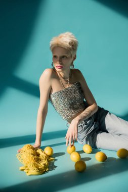 tattooed albino model in shiny top with sequins and jeans sitting near ripe lemons on blue  clipart