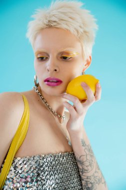 tattooed woman in shiny top with sequins holding ripe lemon on blue  clipart