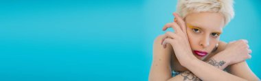 Dövmeli ve parlak makyajlı sarışın albino model mavi arka plandaki kameraya bakıyor, afiş. 
