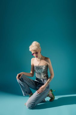 Moda gözlüklü ve mavi arka planda poz veren dövmeli albino modelinin tam uzunluğu 