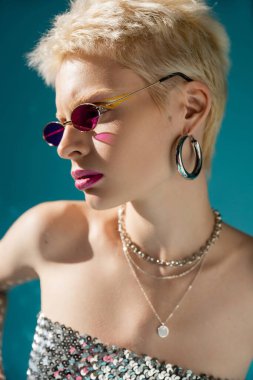 portrait of blonde model in stylish pink sunglasses and jewelry posing on blue  clipart