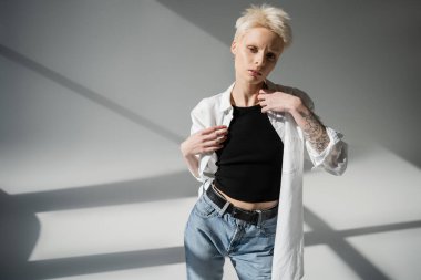 Elinde dövmesi olan genç albino kadın gri arka planda gölgeli beyaz gömlek giyiyor.