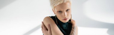 Elinde dövmesi olan genç bir albino kadın. Gri arka planda, gölgeli, pankartıyla kendine sarılıyor. 