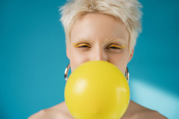 blonde albino woman with bright eye liner blowing bubble gum on blue background 