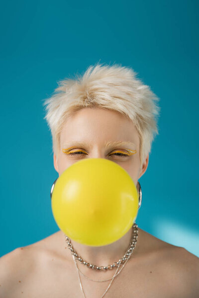 blonde albino woman with yellow eye liner blowing bubble gum on blue background 
