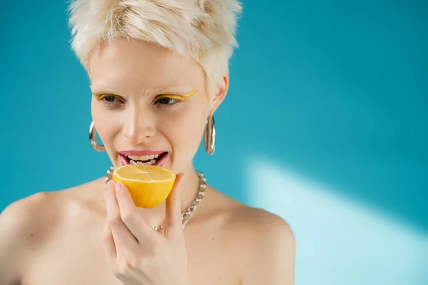 blonde albino woman with bare shoulders biting sour lemon half on blue background
