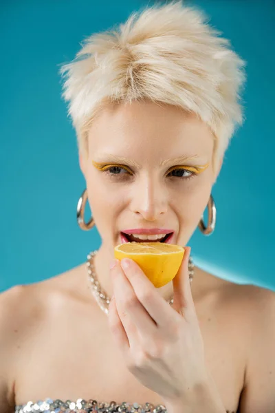blonde albino model with bare shoulders biting sour lemon half on blue background