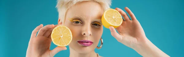 blonde albino model holding sour lemon halves near face isolated on blue, banner