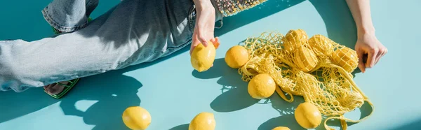 stock image high angle view of young woman in shiny top with sequins and jeans sitting near string bag and ripe lemons on blue, banner 