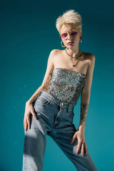 tattooed albino model in trendy pink sunglasses and fashionable outfit posing on blue