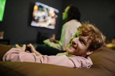 KYIV, UKRAINE - 13 Şubat 2023: Oyun kulübünde bulanık bir arkadaşla video oyunu oynarken kameraya bakan gülümseyen kızıl adam 