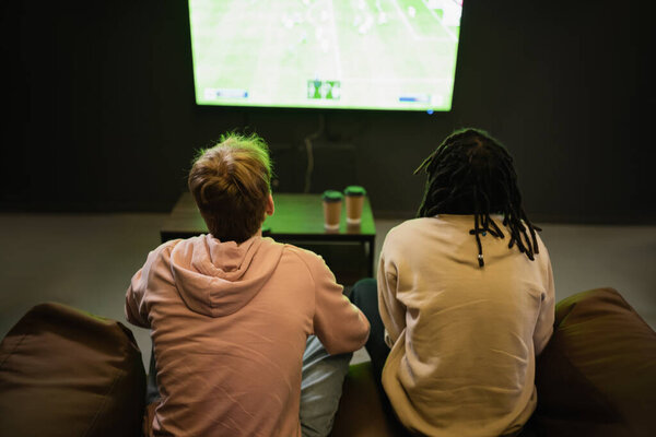 Вид сзади на межрасовых друзей, смотрящих спортивный матч рядом с размытым кофе, чтобы пойти в киберклуб 