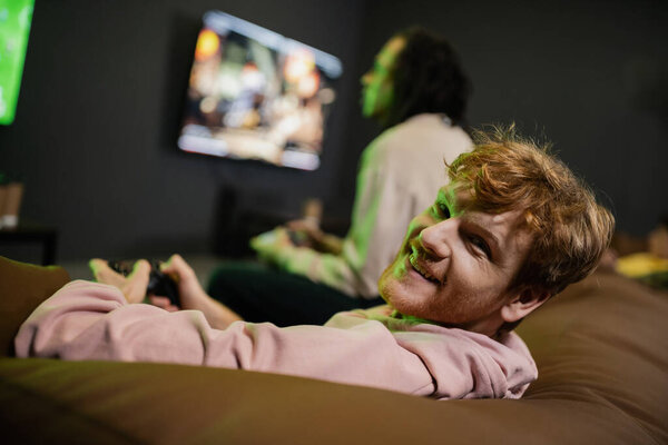 KYIV, UKRAINE - FEBRUARY 13, 2023: Smiling redhead man looking at camera while playing video game with blurred friend in gaming club 