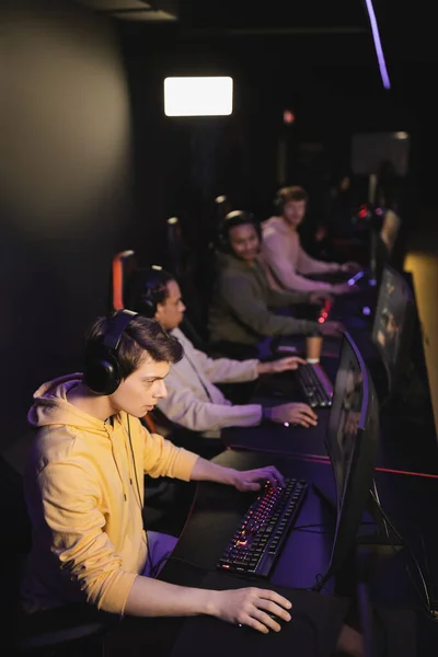 ゲームクラブでぼやけた異人種間のチームの近くにコンピュータ上でビデオゲームをプレイヘッドフォンでゲーマー — ストック写真