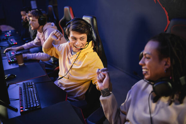 Cheerful man in headphones doing fist bump with multiracial friend in gaming club 