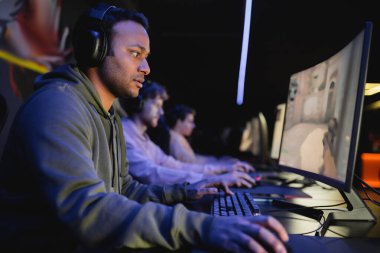 Indian man in headphones playing video game on computer with team in cyber club 